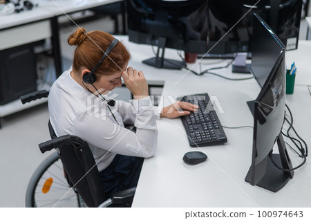 Caucasian woman in a wheelchair sits at her desk and holds her head. Female call center worker. Caucasian woman in a wheelchair sits at her desk and holds her head. Female call center worker. 100974643