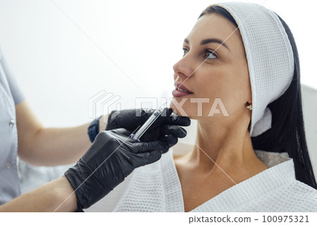 Female cosmetologist in transparent gloves makes an injection in the lower jaw of her client 100975321