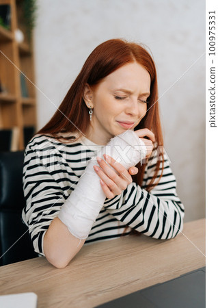 Vertical portrait of suffering from pain crying young woman with broken right hand wrapped in gypsum bandage sitting at table closed eyes. Sad redhead female with broken arm from accident at home. Vertical portrait of suffering from pain crying young woman with broken right hand wrapped in gypsum bandage sitting at table closed eyes. Sad redhead female with broken arm from accident at home. 100975331