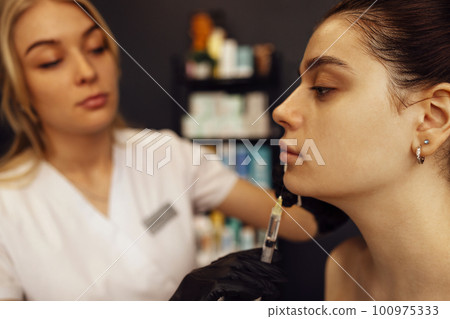 Female cosmetologist in transparent gloves makes an injection in the lower jaw of her client Female cosmetologist in transparent gloves makes an injection in the lower jaw of her client 100975333