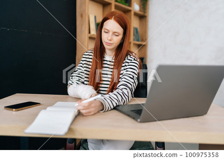 Portrait of successful young businesswoman with broken right hand wrapped in white gypsum bandage working remotely with paper documents and laptop computer, sitting at table from home office room. 100975358