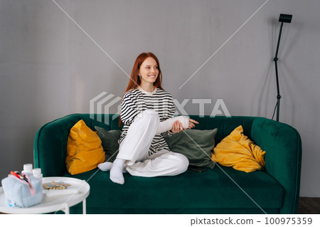Portrait of cheerful young woman with broken hand wrapped in white plaster bandage sitting on sofa in living room smiling looking away. Positive redhead female with broken arm from accident at home. 100975359