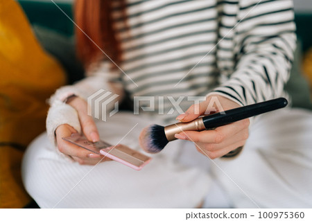Closeup cropped shot of unrecognizable injured young woman with broken arm wrapped in white gypsum bandage applying cosmetic with makeup brush at home. Concept of insurance and healthcare 100975360