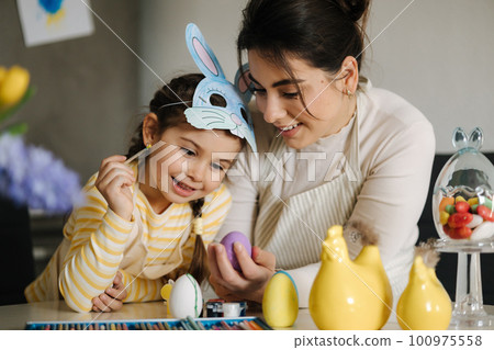 Easter time with family. Adorable girl wore bunny mask and help mom painting eggs. Spring decoration in kitchen Easter time with family. Adorable girl wore bunny mask and help mom painting eggs. Spring decoration in kitchen 100975558