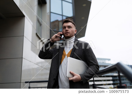 successful european young businessman talking on the phone holding a laptop in his hands on the successful european young businessman talking on the phone holding a laptop in his hands on the 100975573