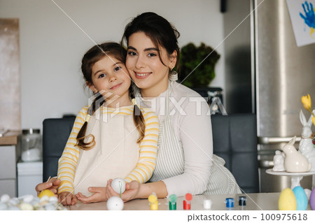 Portrait of adorable little girl with mom on kitchen. Beautiful woman help her daughter paint easter eggs 100975618