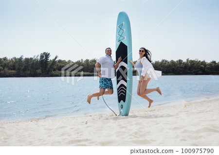Surfers couple on the beach. Man stand with paddleboard and smiling. 100975790