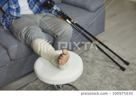 Young African American man with a broken leg sitting with crutches on the sofa at home 100975830