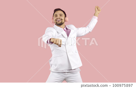 Positive excited handsome guy in white suit and headphones having fun on pink background. 100975897