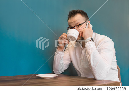 Stylish young guy businessman from Europe drinks coffee and talks on his smartphone sitting at a table in a cafe on a break 100976339