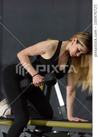 Athlete woman in sportswear training back with dumbbell in one hand leaning on the bench 100976725