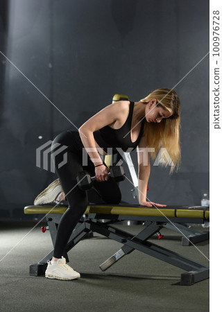 Athlete woman in sportswear training back with dumbbell in one hand leaning on the bench 100976728