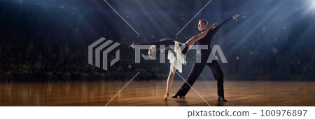 Dance aesthetics. Talented young man and girl in beautiful costumes performing, dancing tango and ballroom dance under spotlights. 3D audience background Dance aesthetics. Talented young man and girl in beautiful costumes performing, dancing tango and ballroom dance under spotlights. 3D audience background 100976897