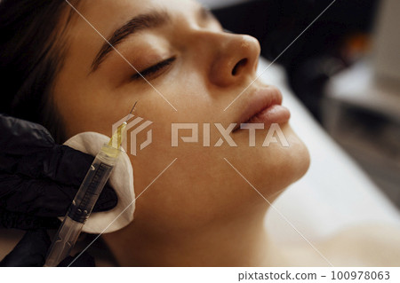 Female cosmetologist in transparent gloves makes an injection in the lower jaw of her client 100978063