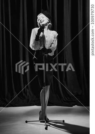 Adorable woman wearing vintage clothes standing with microphone and singing. Monochrome Adorable woman wearing vintage clothes standing with microphone and singing. Monochrome 100979730