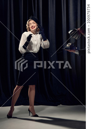 Adorable woman wearing vintage clothes holding phone and talking with smiling over dark background Adorable woman wearing vintage clothes holding phone and talking with smiling over dark background 100979734