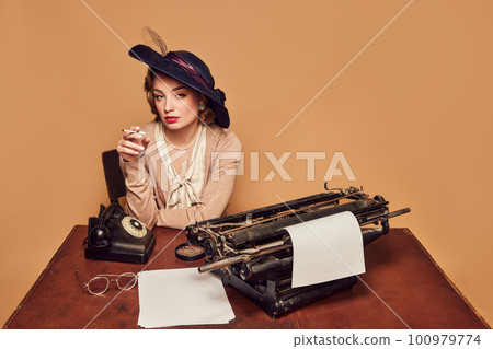 Attractive woman writer wearing old-fashioned clothes sitting at table and smoking over beige background 100979774
