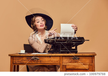 Surprised beautiful woman writer wearing retro clothes sitting at typewriter with astonished face over beige background 100979788