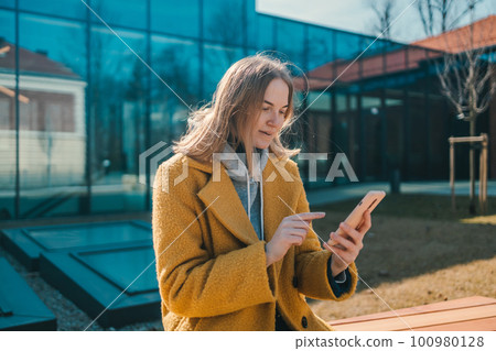 Photo portrait of blonde happy student woman 20s holding phone wearing a yellow coat and sitting on bench in city spring park outdoors resting use mobile cell phone chat online. 100980128