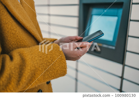 Self service post terminal machine and parcel sending. Woman holding smartphone using red automated self service post terminal machine or locker to deposit the parcel for storage. 100980130