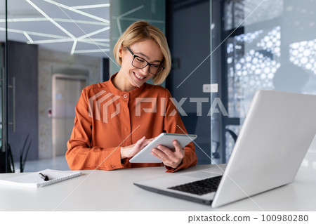 Young successful businesswoman at workplace is satisfied with results of achievement at work smiling and using tablet computer, blonde programmer is testing new software in application. 100980280