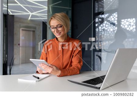Young successful businesswoman at workplace is satisfied with results of achievement at work smiling and using tablet computer, blonde programmer is testing new software in application. 100980282