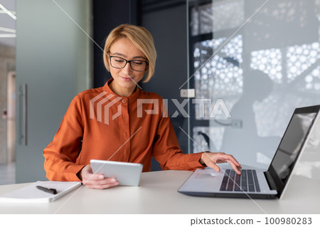 Young successful businesswoman at workplace is satisfied with results of achievement at work smiling and using tablet computer, blonde programmer is testing new software in application. Young successful businesswoman at workplace is satisfied with results of achievement at work smiling and using tablet computer, blonde programmer is testing new software in application. 100980283