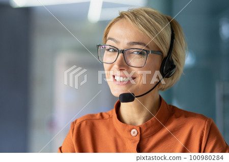 Close-up portrait of young beautiful blonde woman at workplace, call center worker smiling and looking at camera, operator using headset phone for video call and online customer consultation support. 100980284