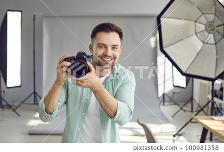 Happy smiling photographer holding a camera standing in professionally studio with light equipment. Happy smiling photographer holding a camera standing in professionally studio with light equipment. 100981358