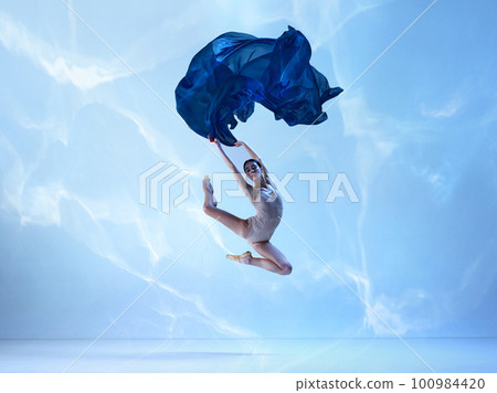 Shot of flexible ballerina, dancer in high jump with navy fabric over blue background. Action, motion, art concept Shot of flexible ballerina, dancer in high jump with navy fabric over blue background. Action, motion, art concept 100984420