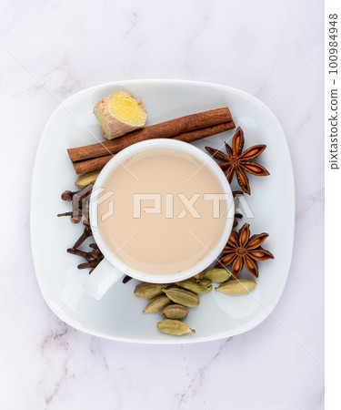 White cup with masala chai on a saucer and cinnamon, anise with cloves and ginger root with cardamom. White cup with masala chai on a saucer and cinnamon, anise with cloves and ginger root with cardamom. 100984948