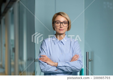 Serious and successful business woman boss inside office building at workplace, woman looking at camera thoughtfully with crossed arms and blue shirt. Serious and successful business woman boss inside office building at workplace, woman looking at camera thoughtfully with crossed arms and blue shirt. 100985703