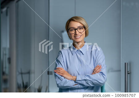 Portrait of young successful business woman inside office at workplace, female worker standing with arms crossed smiling and looking at camera, woman in blue shirt and glasses blonde. Portrait of young successful business woman inside office at workplace, female worker standing with arms crossed smiling and looking at camera, woman in blue shirt and glasses blonde. 100985707