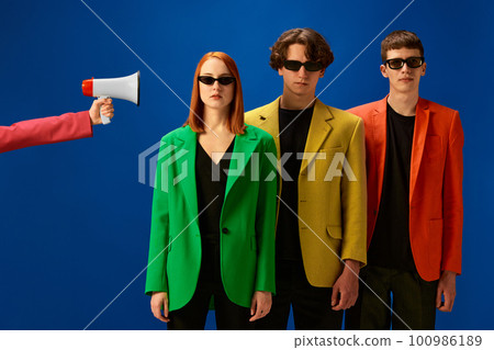 Portrait of young people wearing colorful jackets standing near loudspeaker over blue background Portrait of young people wearing colorful jackets standing near loudspeaker over blue background 100986189