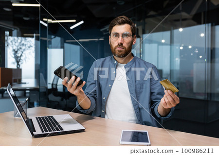 Portrait of disappointed and deceived businessman inside office, mature man looking at camera confused holding bank credit card and phone, employee got rejection error. 100986211