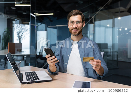 A smiling young man is holding a phone and a credit card. Stands in the office, dials on the phone, makes online purchases, orders, checks the account A smiling young man is holding a phone and a credit card. Stands in the office, dials on the phone, makes online purchases, orders, checks the account 100986212