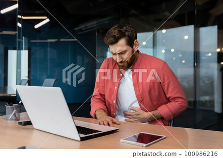 A man in the office at the workplace is sick, he has a severe stomach ache, the businessman got food poisoning from lunch, he is sitting at the table inside the building. A man in the office at the workplace is sick, he has a severe stomach ache, the businessman got food poisoning from lunch, he is sitting at the table inside the building. 100986216