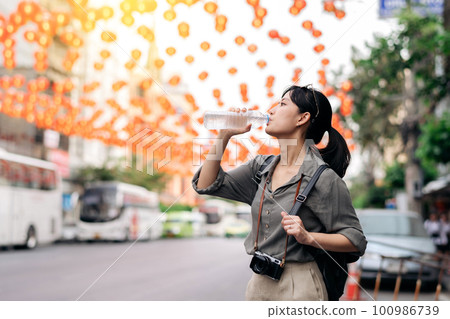 Happy young Asian woman backpack traveler drinking a cold water at China town street food market in Bangkok, Thailand. Traveler checking out side streets. 100986739