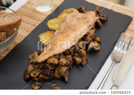 fried trout fillet on pillow of fried champignon mushrooms on dark warm stone plate 100986835