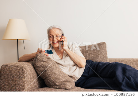 Senior woman looking at credit card talking on a mobile phone. Aged female making online shopping while lying on a sofa.	 100987156
