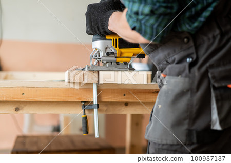 Close-up of a man cutting a wooden plank with an electric jigsaw in a workshop. 100987187