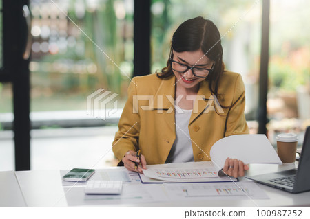 Concept of business office woman working,Businesswoman smile while working about her invesment plan with analyzing document and business investment graph data by using laptop on desk in workstation. 100987252