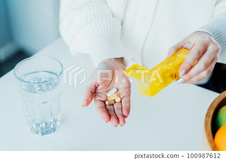 Close-up of woman's hand holding pills and pill organizer at home kitchen with water glass. Daily intake of antioxidant diet, vitamin supplements for beauty, skin, hair, healthcare, drugs concept. 100987612