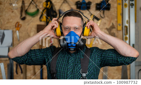 A carpenter in goggles and a respirator puts on noise-protection earmuffs.  100987732