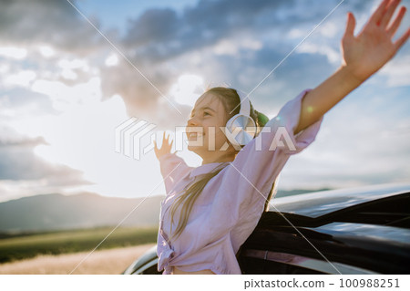 Little girl with headphones standing and leaning out of the car window during the ride. 100988251