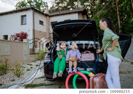 Happy sisters with their mother sitting in car trunk and waiting for electric car charging. 100988255