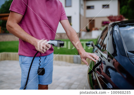 Man holding power supply cable at electric vehicle charging station. 100988285