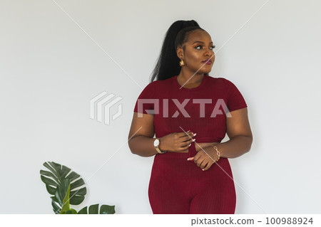 Portrait of fat young african american woman standing against white wall indoors copy space and empty space for text. Female wearing a red short t-shirt and leggings. Sports home wear and inclusion Portrait of fat young african american woman standing against white wall indoors copy space and empty space for text. Female wearing a red short t-shirt and leggings. Sports home wear and inclusion 100988924
