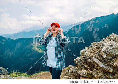 woman with backpack meditating listen to music with headphones and looking view landscape river peak mountain, female tourist with long blond hair relax after walking enjoy journey trip, copy space 100989672