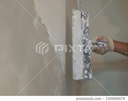 gloved hand of a plasterer holding a wide trowel against a freshly plastered empty wall, fragment of wall finishing work, hand construction tool in builder's hand 100990079